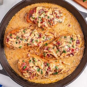 Three cooked chicken breasts in a creamy sauce with onions and herbs, this smothered chicken is served hot in a black skillet atop a marble surface.