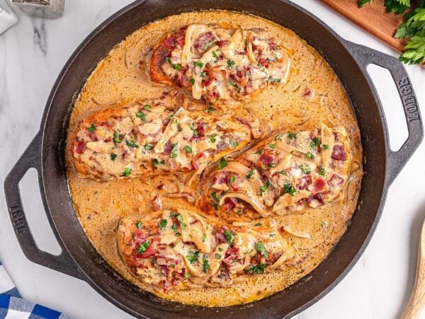 Three cooked chicken breasts in a creamy sauce with onions and herbs, this smothered chicken is served hot in a black skillet atop a marble surface.