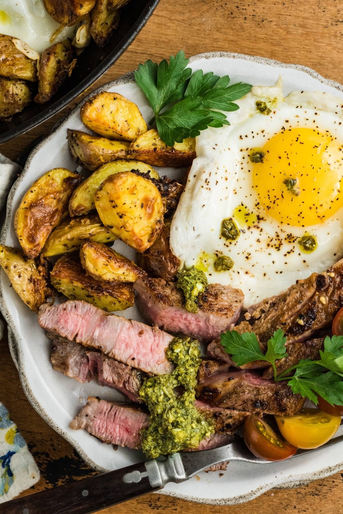 A plate with sliced steak, roasted potatoes, a sunny-side-up egg, cherry tomatoes, and herbs.