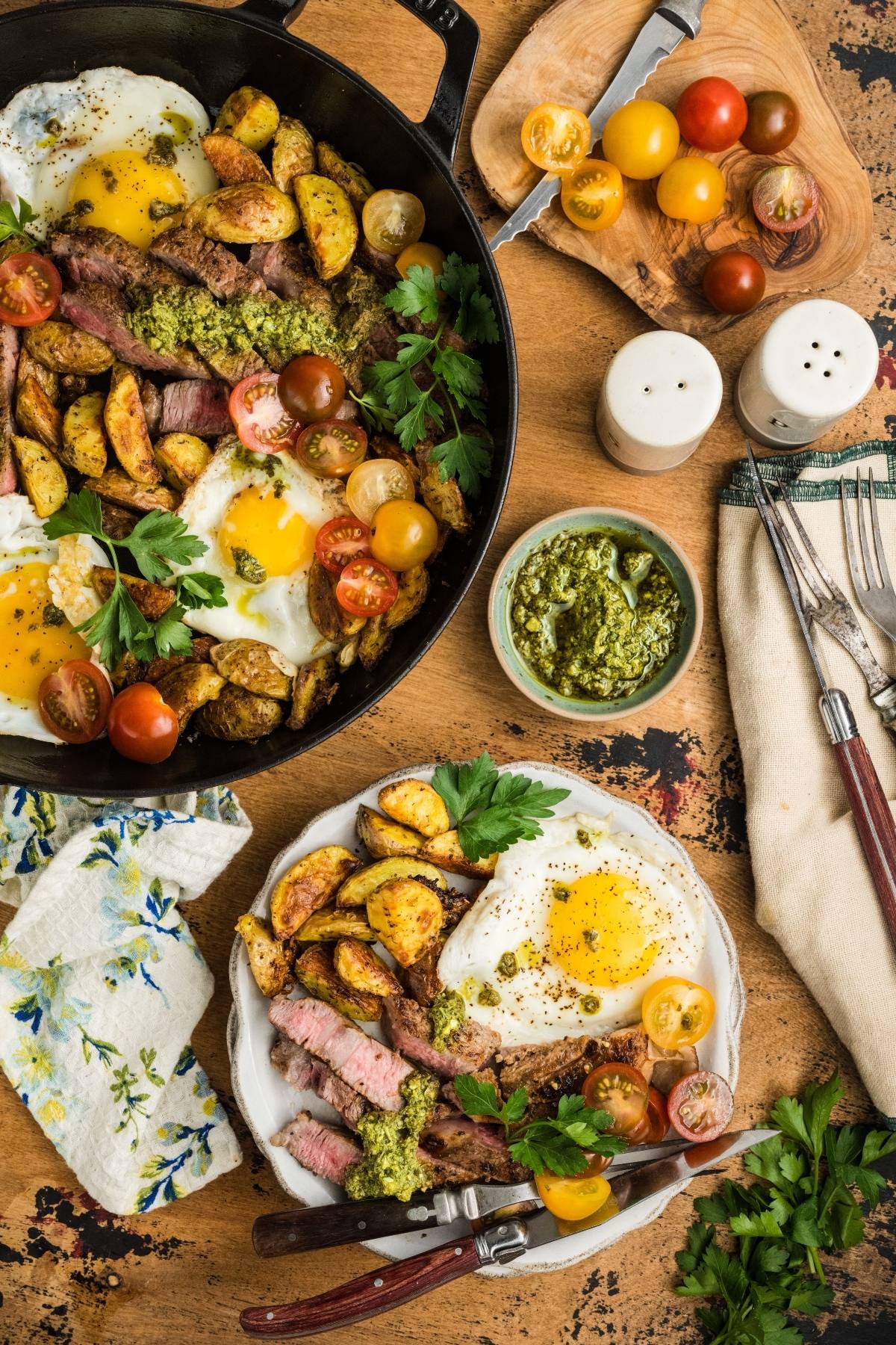 Pan and plate with steak, fried eggs, roasted potatoes, cherry tomatoes, pesto, and fresh parsley on a rustic table.