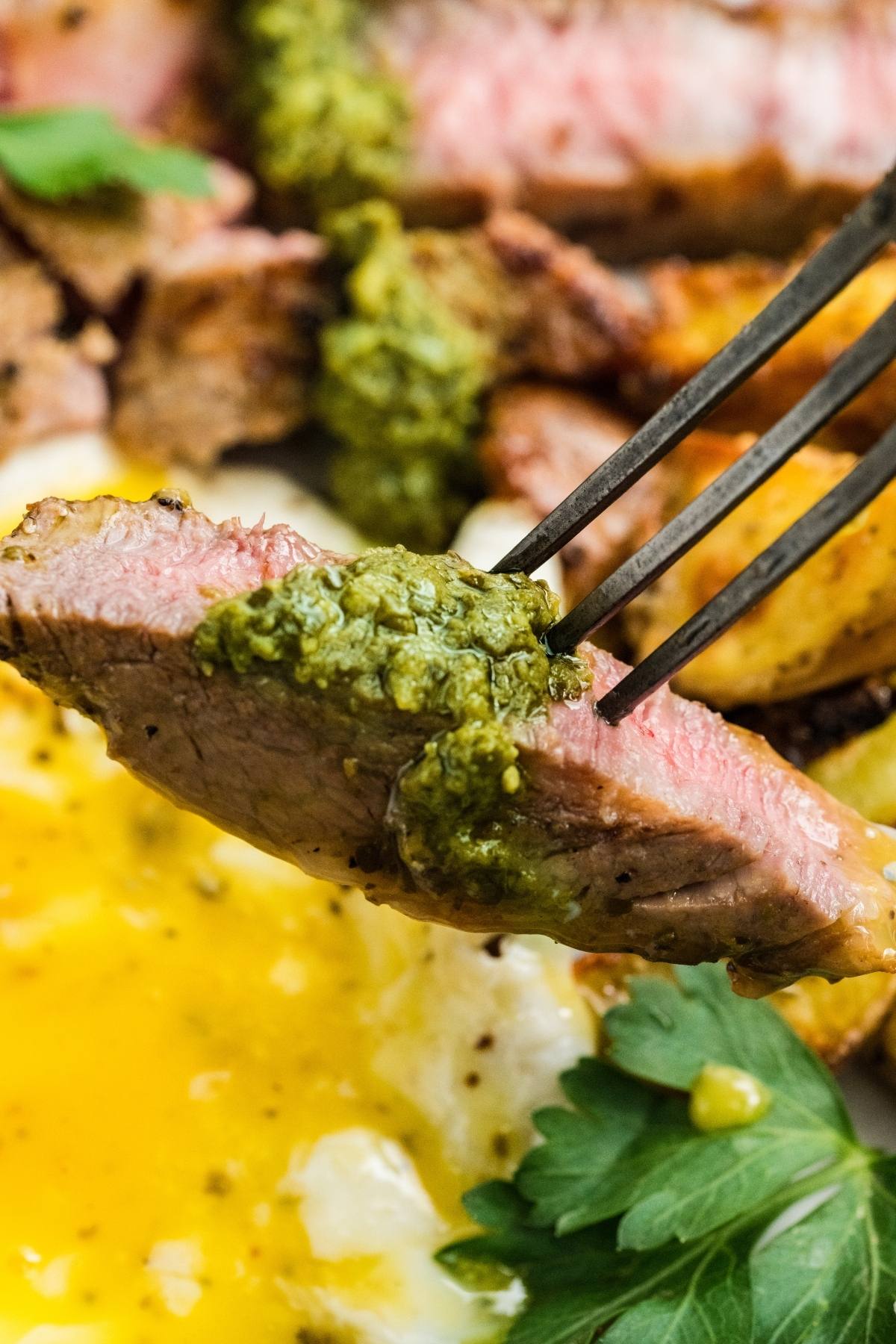 A fork holds a slice of steak with green sauce, above eggs with runny yolk and fresh parsley.