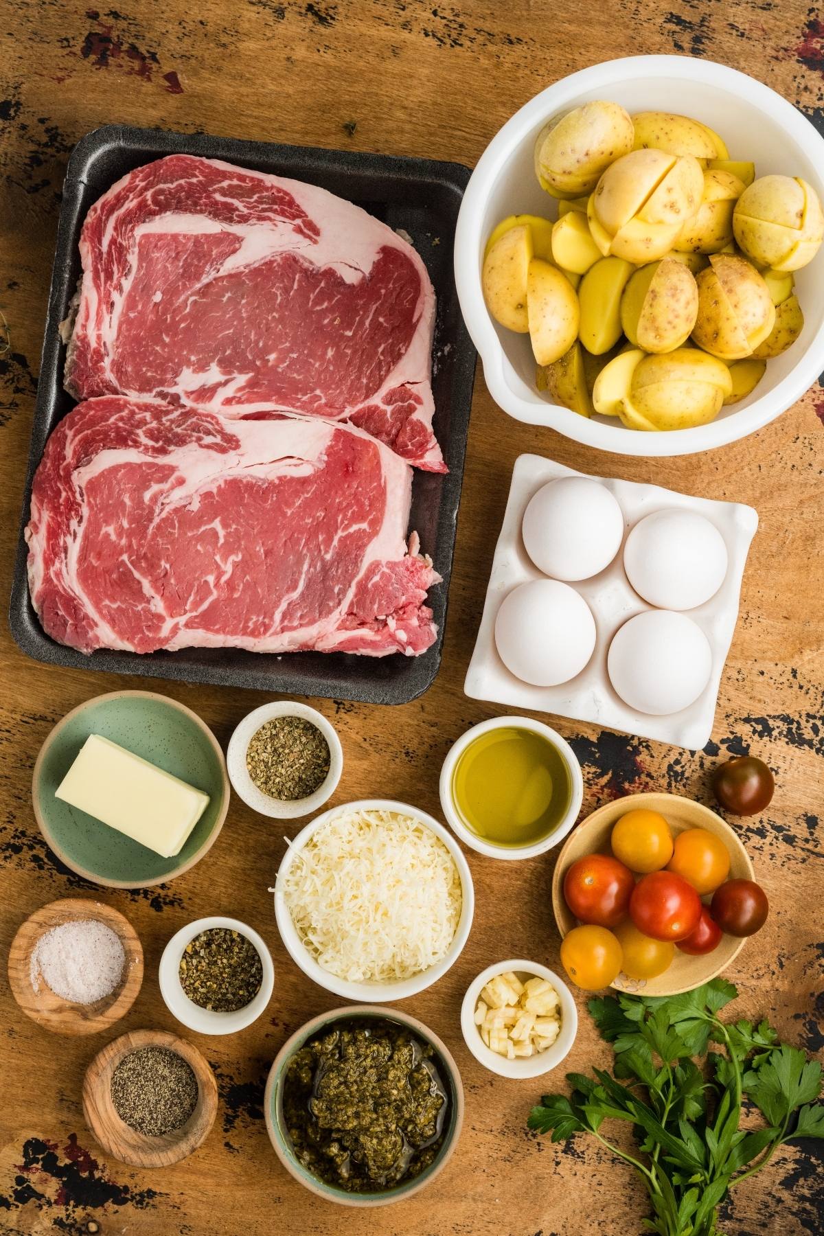 Raw ribeye steaks, halved potatoes, eggs, cherry tomatoes, butter, herbs, cheese, oil, and seasonings on a table.