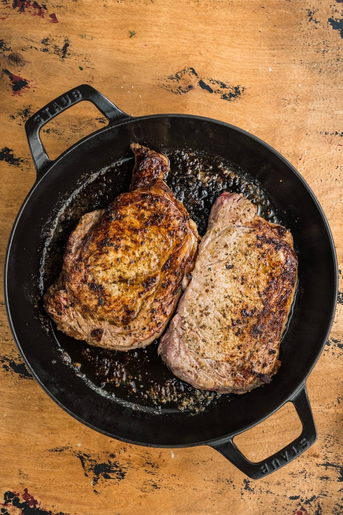 Two seared steaks cooking in a black cast iron skillet on a wooden surface.