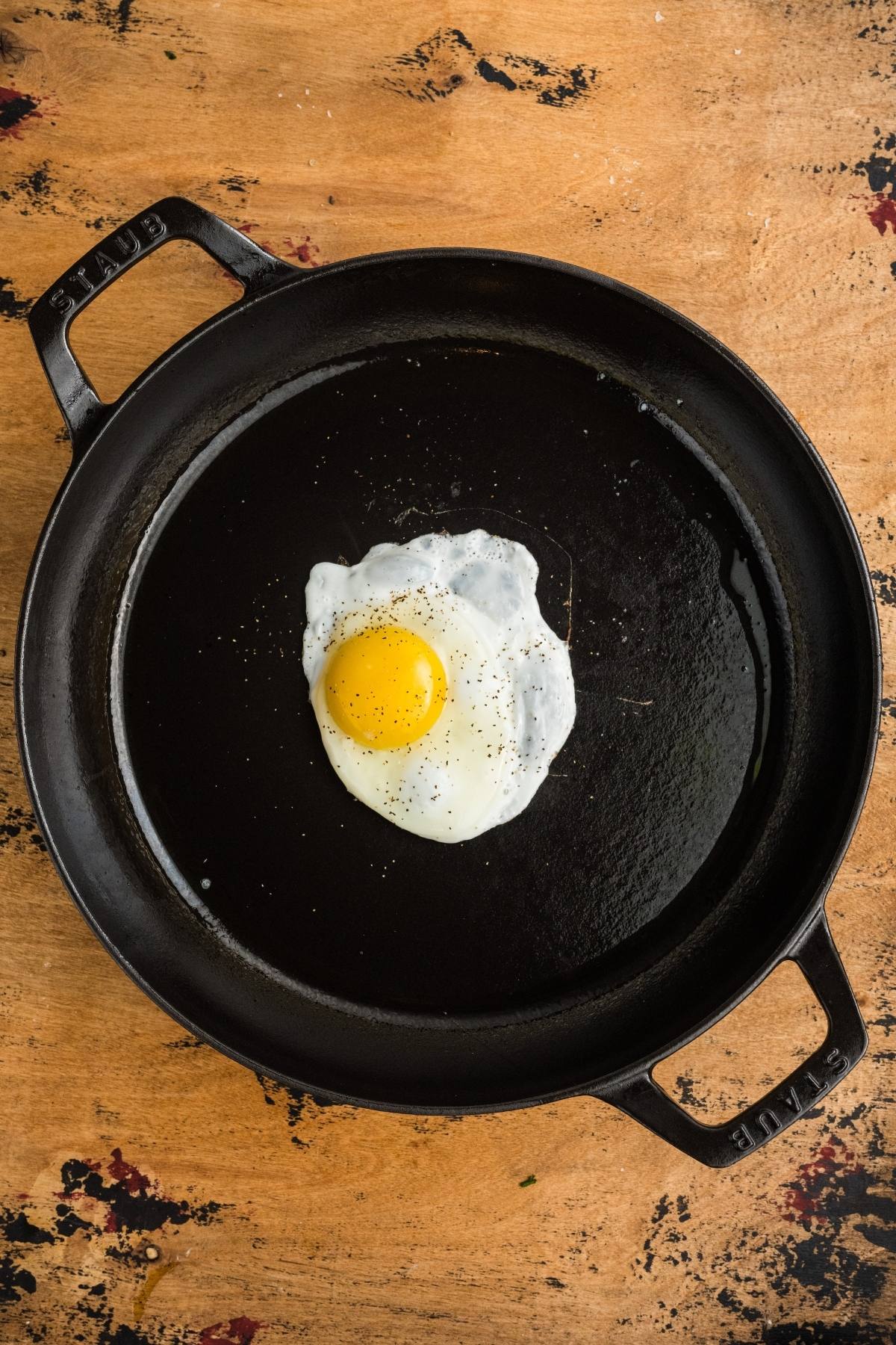 A single fried egg with pepper in a black cast iron skillet on a wooden surface.