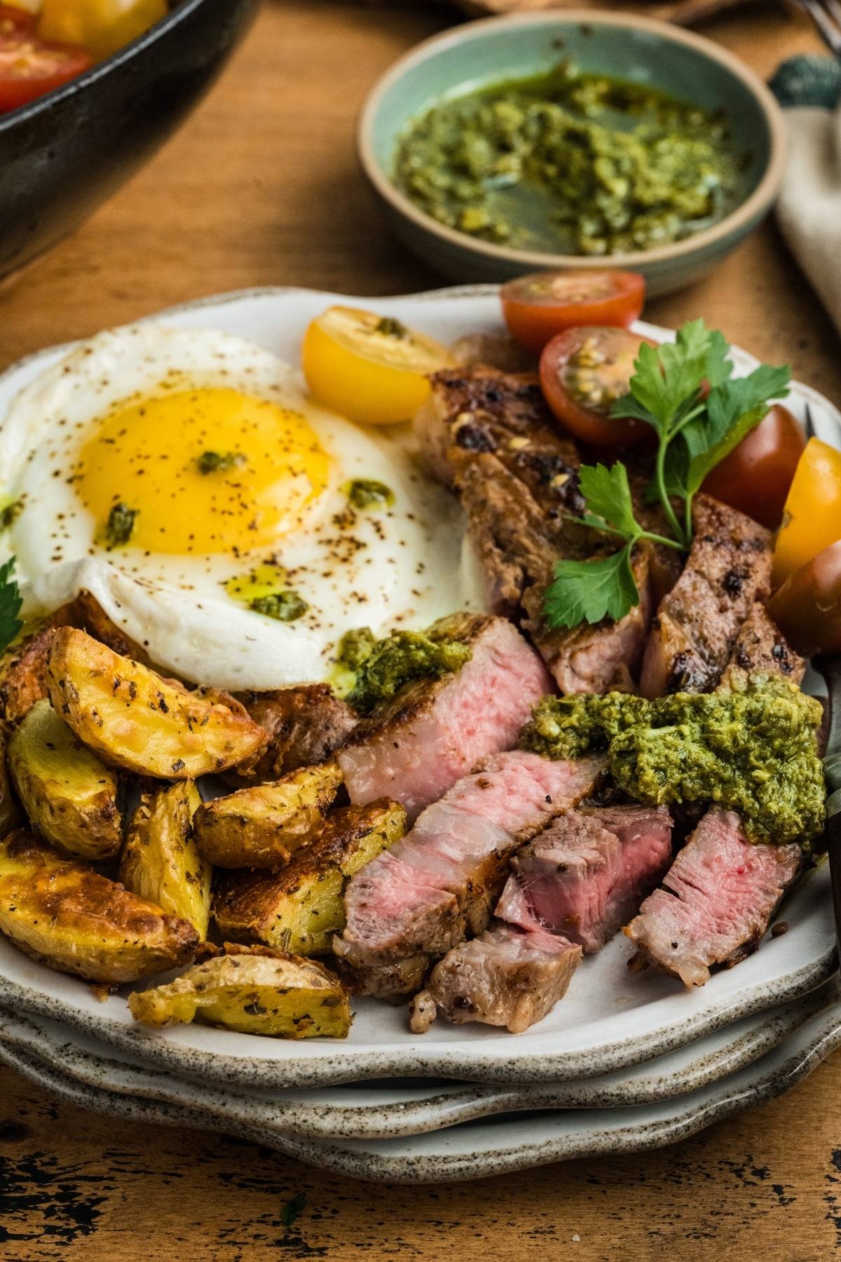 A plate with steak slices, roasted potatoes, a sunny-side up egg, tomatoes, pesto, and parsley garnish.