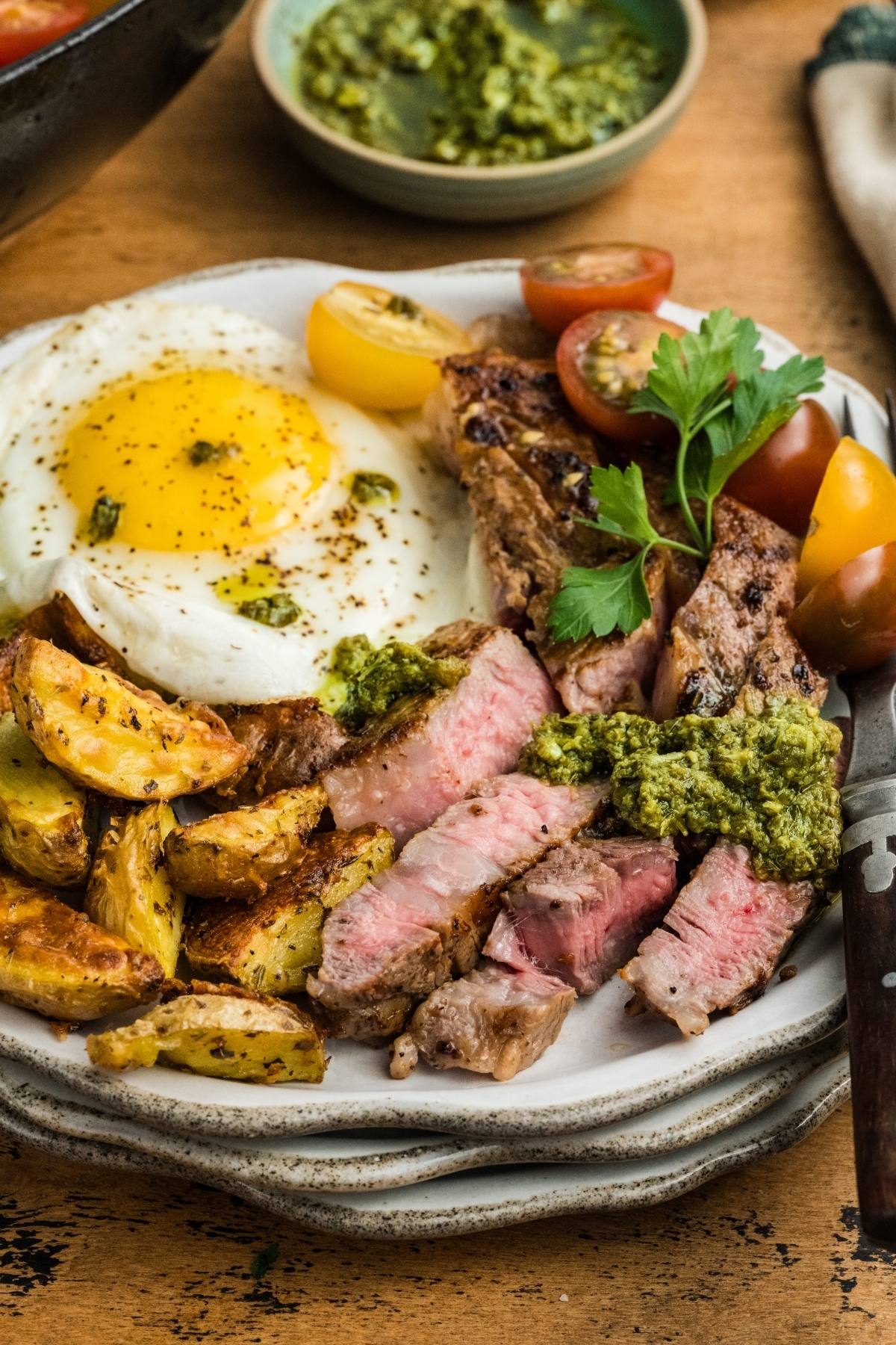 Plate with sliced steak, fried egg, roasted potatoes, cherry tomatoes, and green herb sauce.