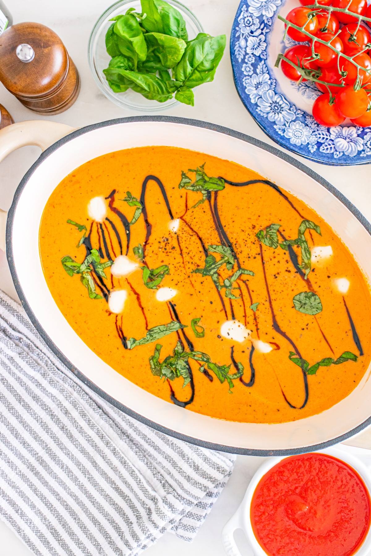 Oval dish of creamy tomato soup garnished with basil, cream, and balsamic drizzle, surrounded by fresh ingredients.