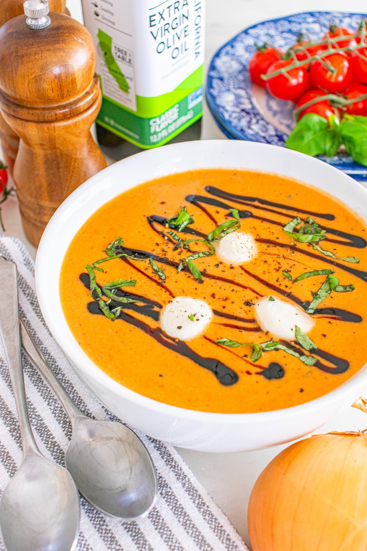 Bowl of tomato soup with cream, basil, and balsamic drizzle, next to spoons, olive oil, and fresh tomatoes.