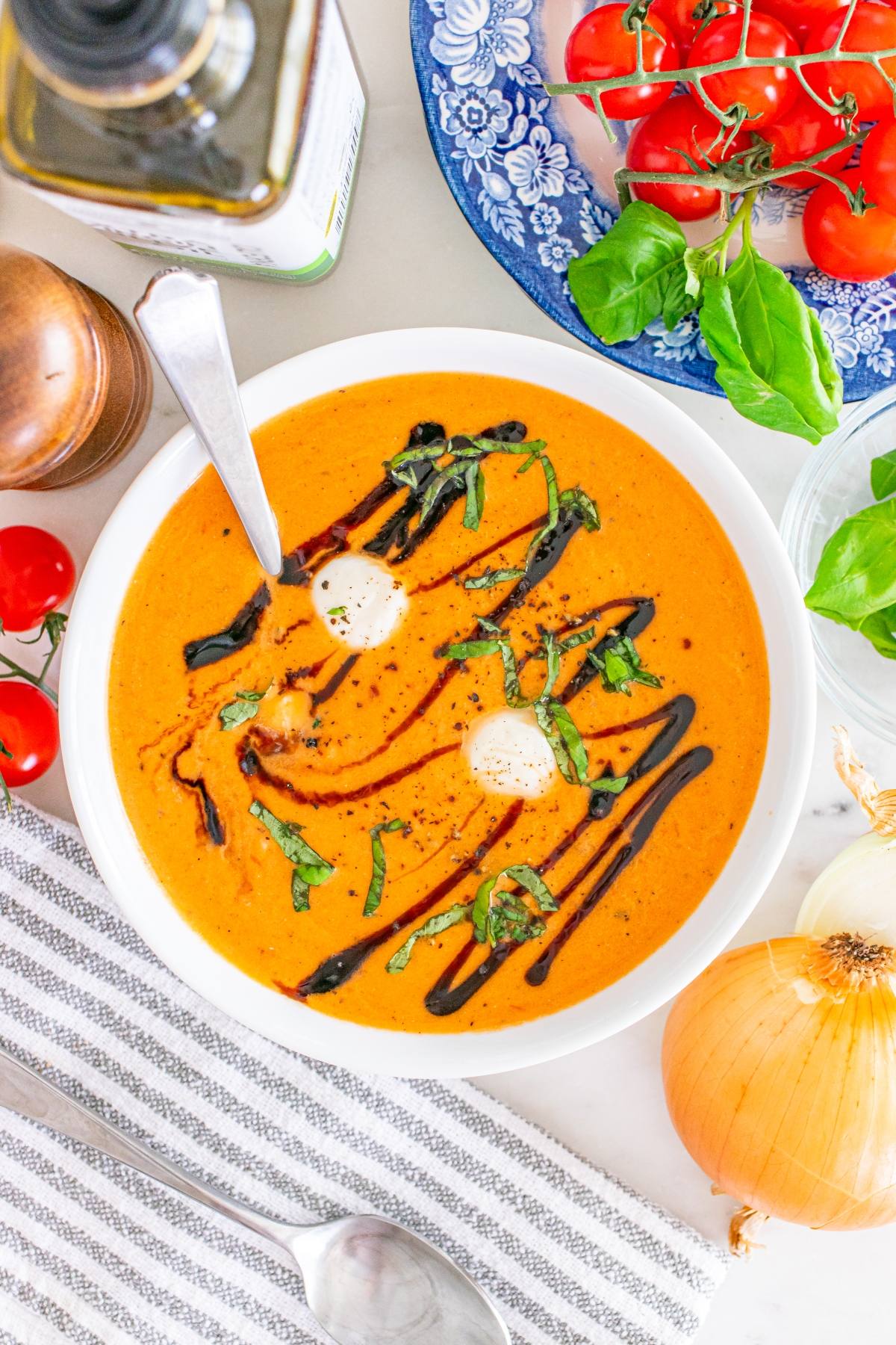 A bowl of tomato soup with cream, basil, and balsamic drizzle, surrounded by fresh tomatoes and basil.