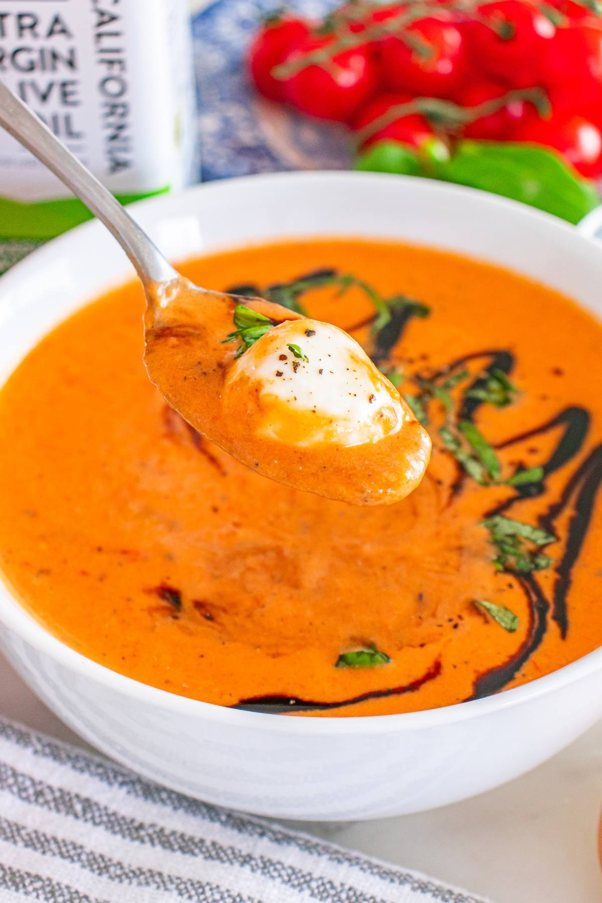 Spoonful of creamy tomato soup with burrata cheese and herbs above a bowl of soup, garnished with basil oil.