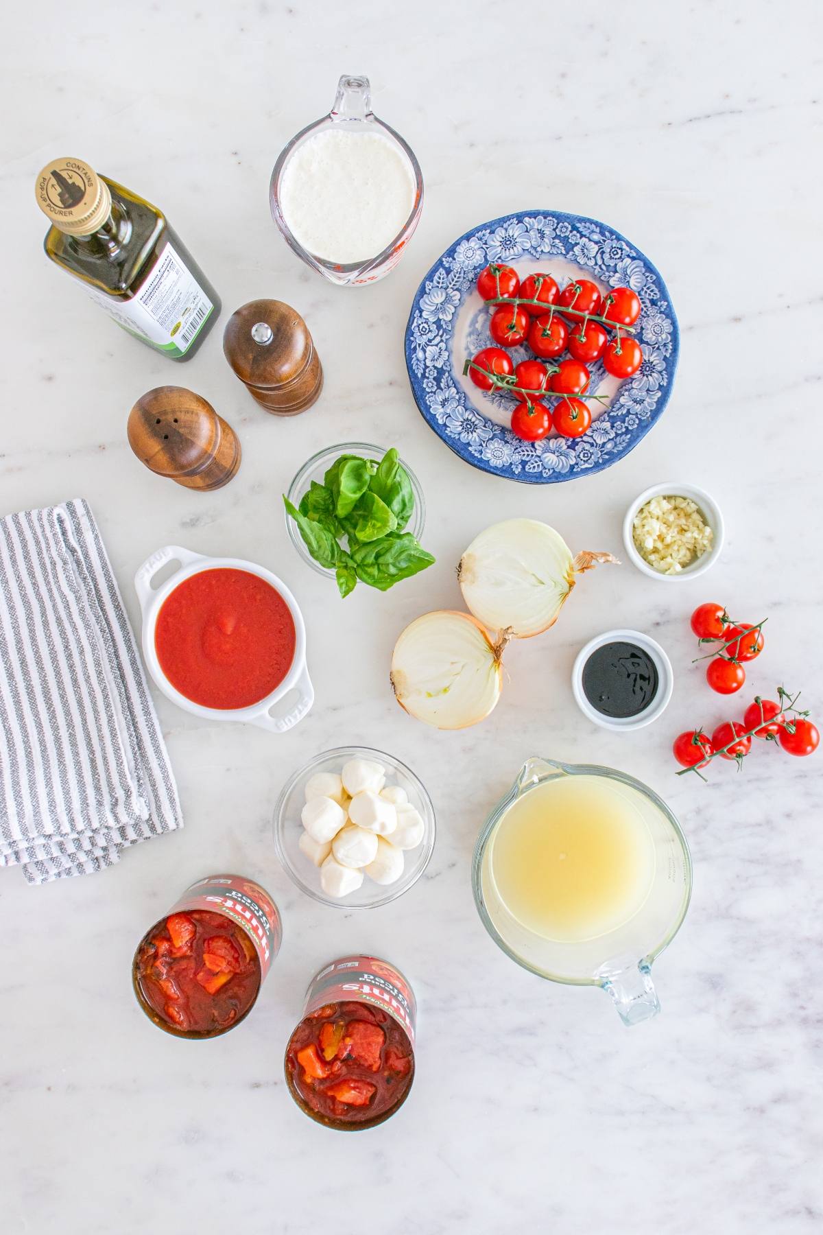 Ingredients for tomato soup on a marble countertop, including tomatoes, onions, basil, cheese, broth, and seasonings.