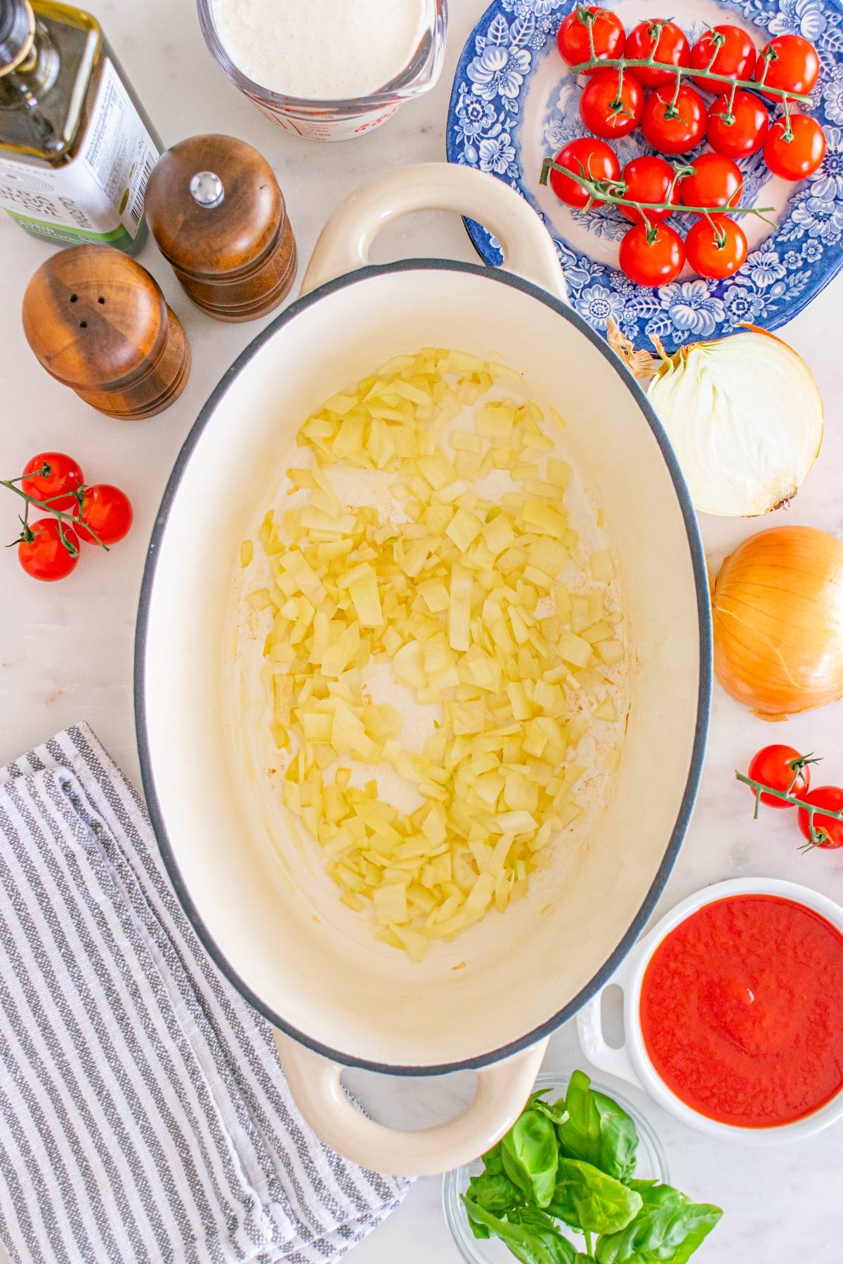 Chopped onions cooking in a large pot, surrounded by tomatoes, basil, seasonings, and a striped towel.
