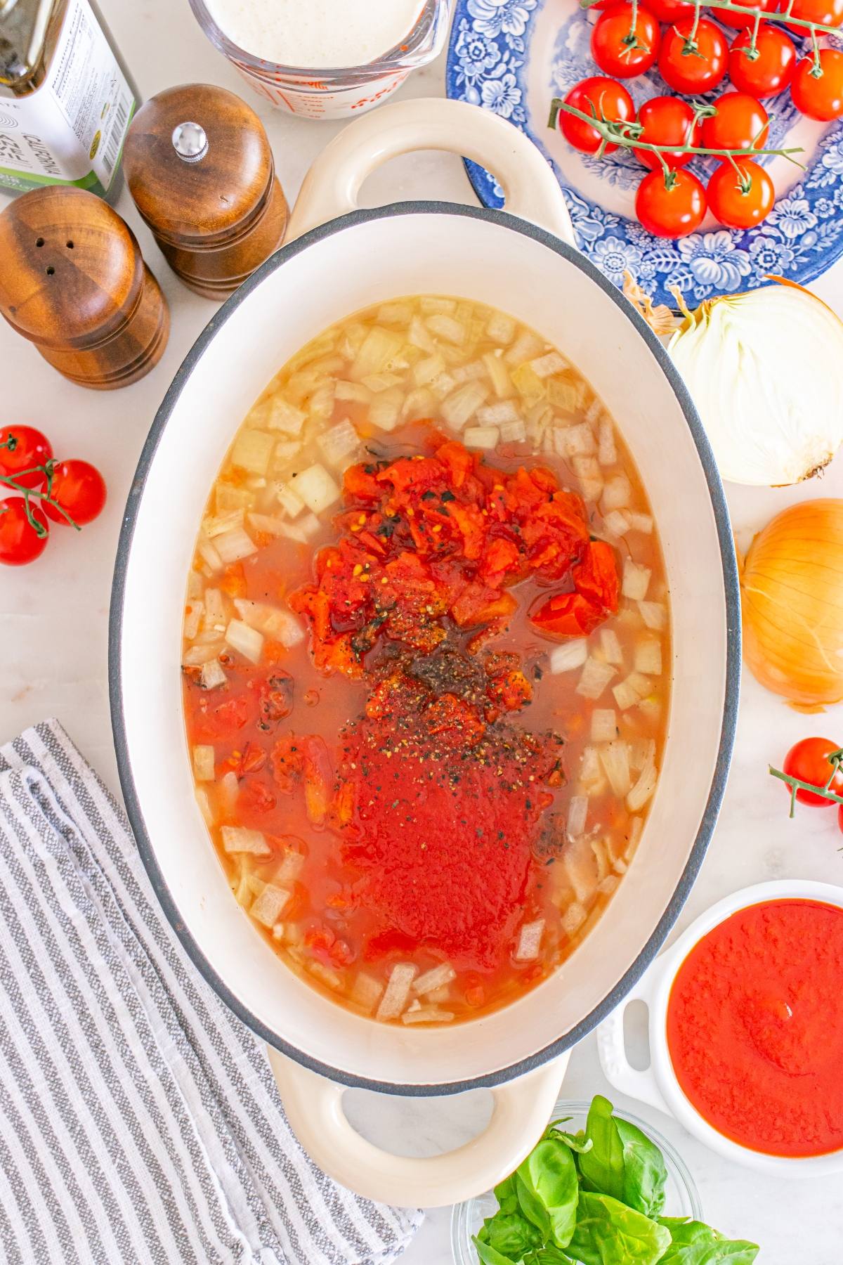 Oval pot with chopped onions, diced tomatoes, and seasonings; fresh tomatoes and basil nearby on the counter.