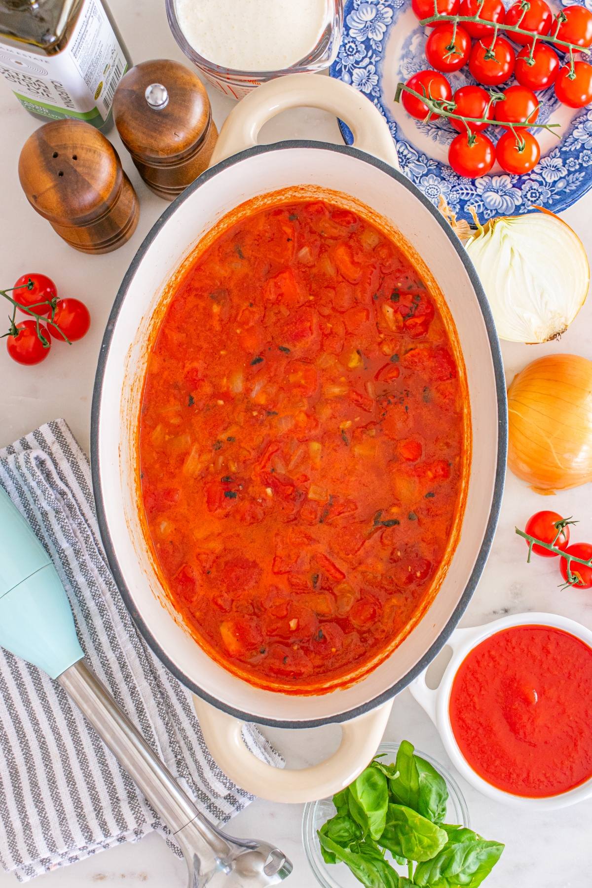 A pot of tomato sauce surrounded by fresh ingredients like tomatoes, onion, basil, and kitchen utensils.