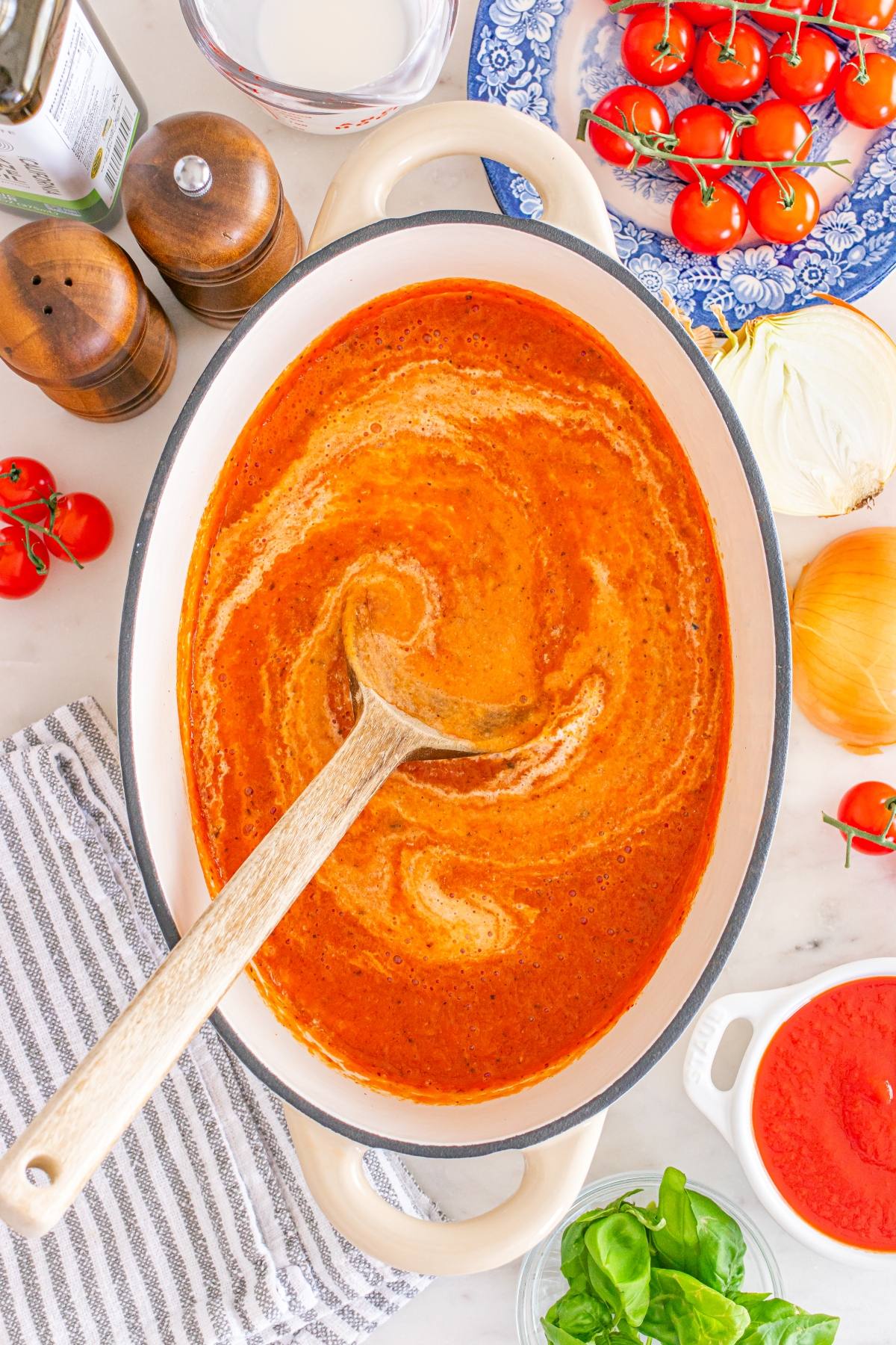 Wooden spoon stirring creamy tomato soup in a pot, surrounded by tomatoes, onion, basil, and seasonings.