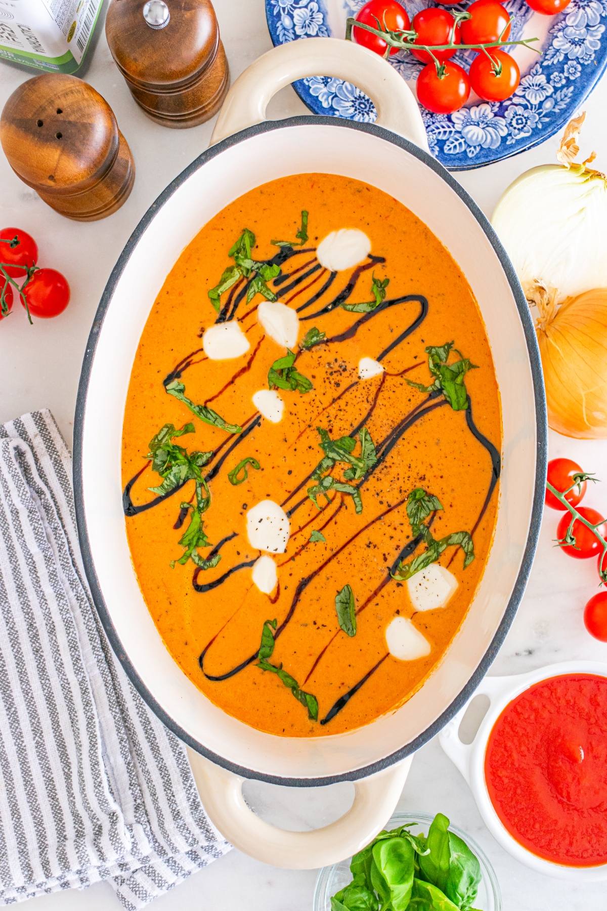 Oval pot of creamy tomato soup, garnished with cream, basil, and balsamic glaze, surrounded by fresh ingredients.