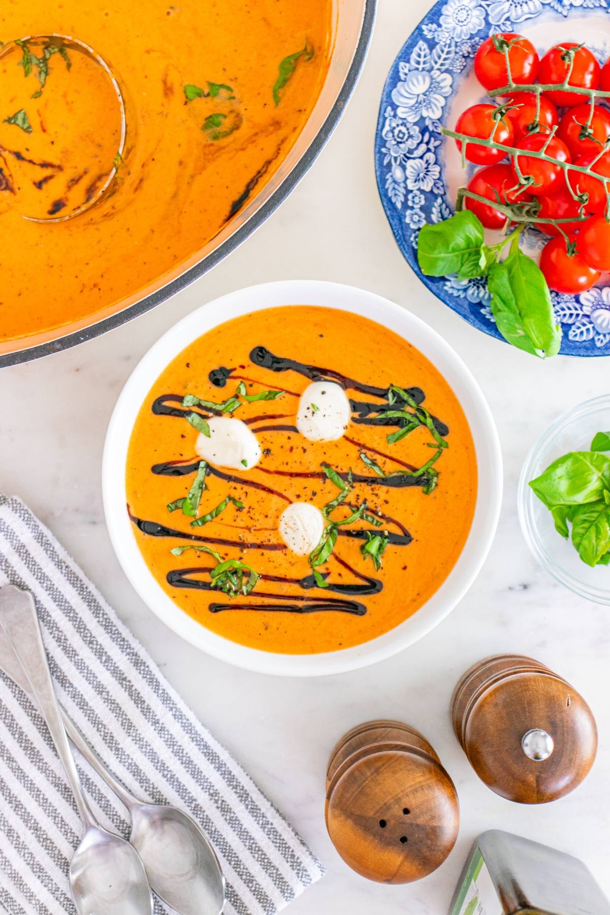 Bowl of tomato soup with cream, basil, and balsamic drizzle, next to fresh tomatoes and basil.
