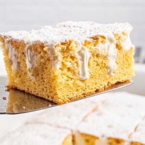 A square slice of Twinkie Poke Cake with white icing and powdered sugar sits on a spatula above a baking dish.