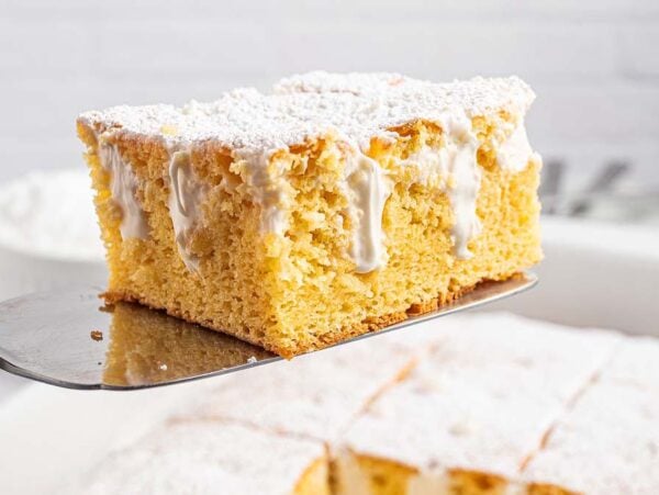 A square slice of Twinkie Poke Cake with white icing and powdered sugar sits on a spatula above a baking dish.