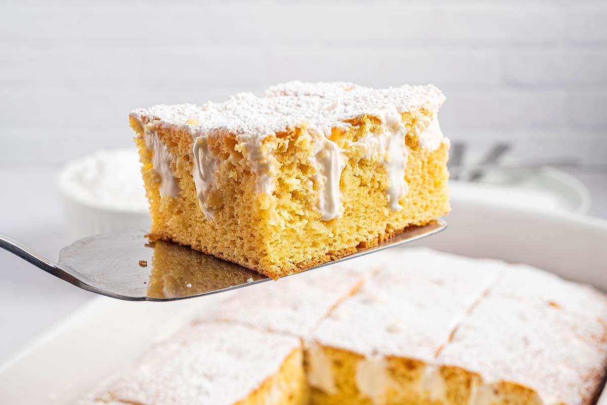 A spatula lifts a slice of Twinkie Poke Cake, topped with white icing and powdered sugar, from a baking dish.