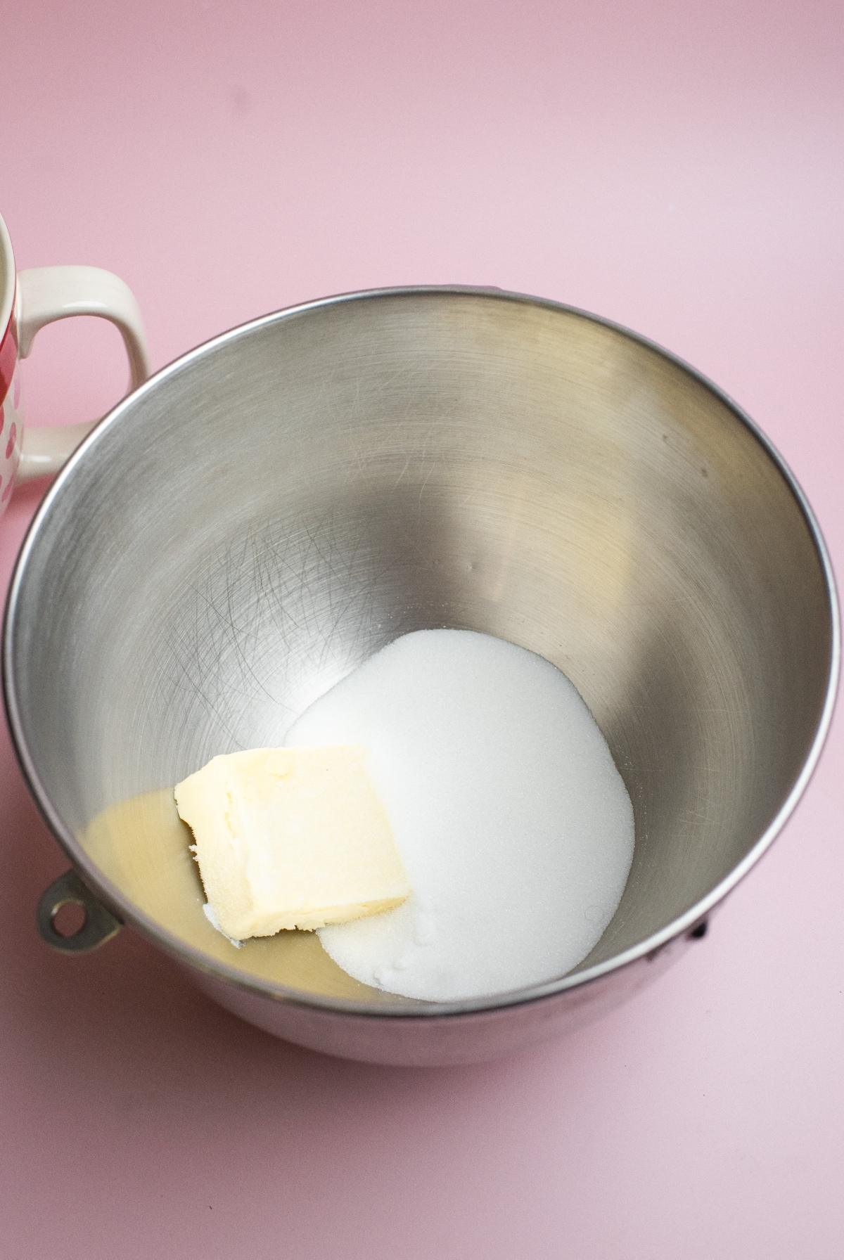 A metal bowl with butter and sugar inside, on a pink surface next to a mug—perfect for making Valentine’s Day sugar cookies or Cream Cheese Sprinkle Dip.