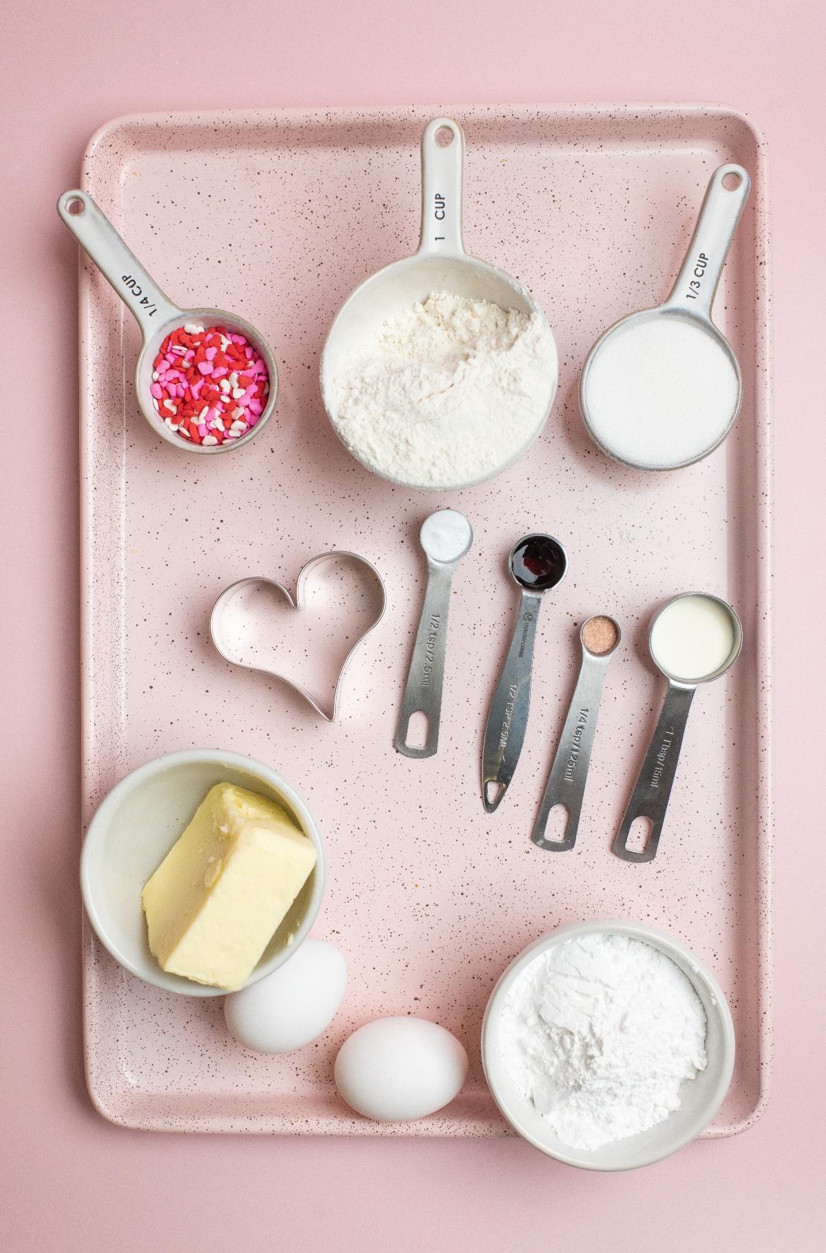 Baking ingredients on a pink, heart-shaped tray: flour, sugar, butter, eggs, sprinkles, and measuring spoons with liquids—perfect for whipping up Valentine’s party treats like Cream Cheese Sprinkle Dip.