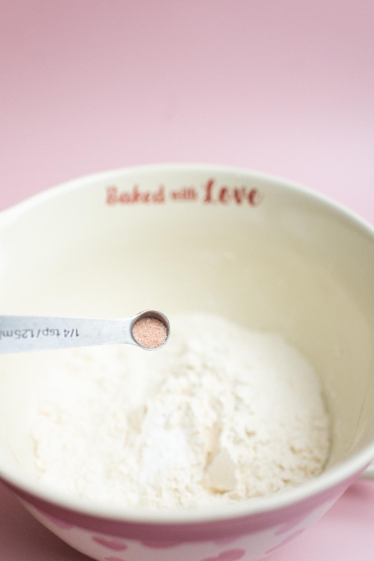 A measuring spoon of salt held over a bowl of flour on a pink background—perfect for whipping up Valentine’s day sugar cookies or other sweet Valentine’s party treats.