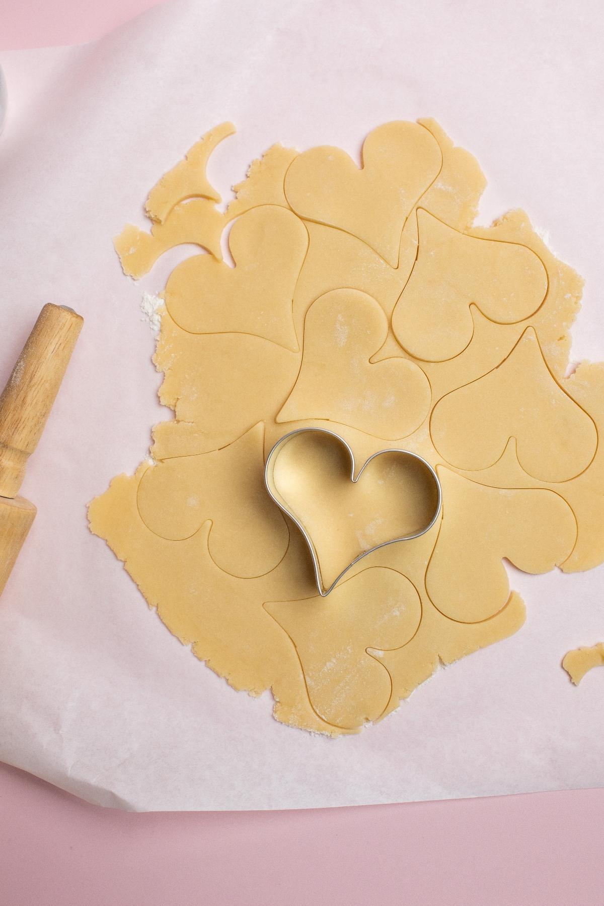 A heart shaped cookie cutter presses into rolled dough for charming Valentine’s party treats, with heart shapes on parchment paper and a rolling pin nearby.