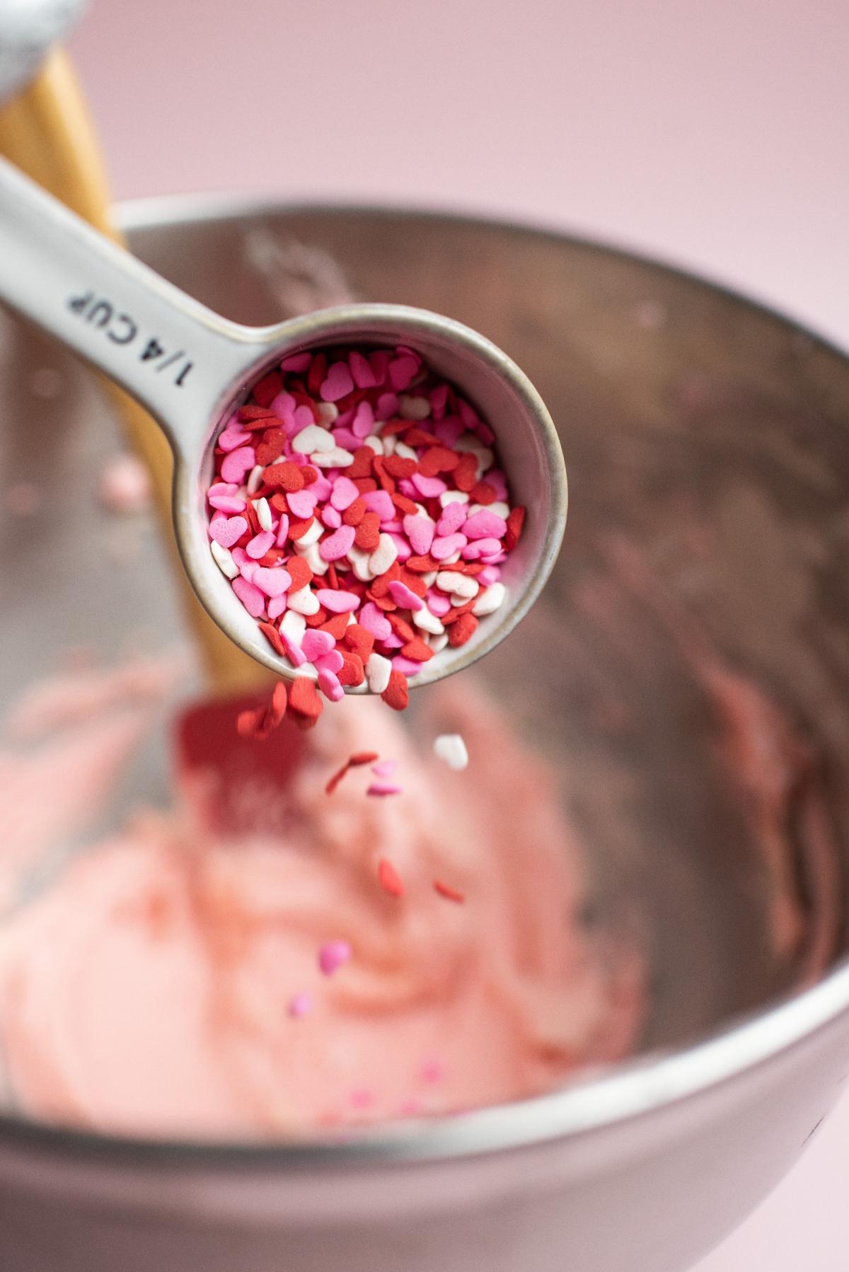 A measuring cup of heart shaped pink sprinkles held over a bowl of pink frosting, perfect for decorating Valentine’s day sugar cookies or festive Valentine’s party treats.