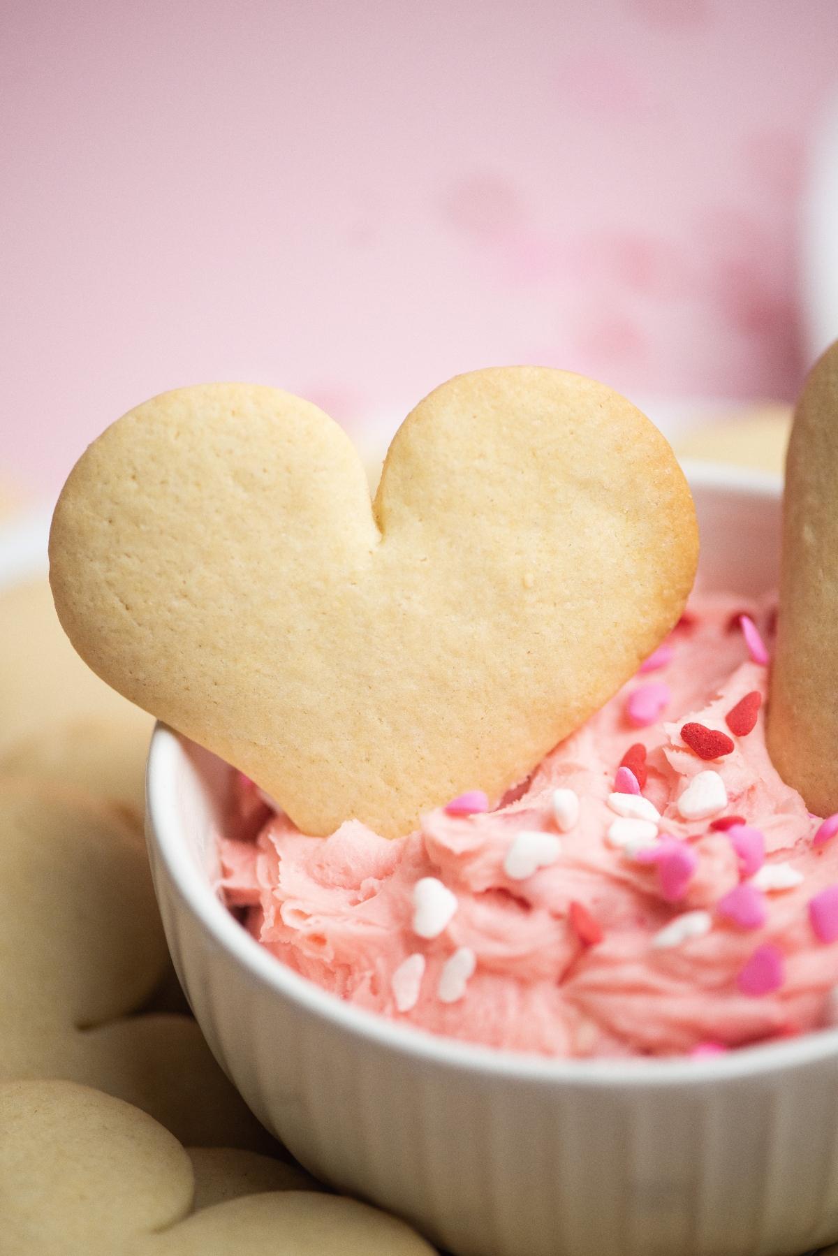 Heart-shaped cookie in pink frosting with heart-shaped sprinkles, in a white bowl—perfect for serving alongside Valentine Cookies and Dip at your Valentine’s party treats table.