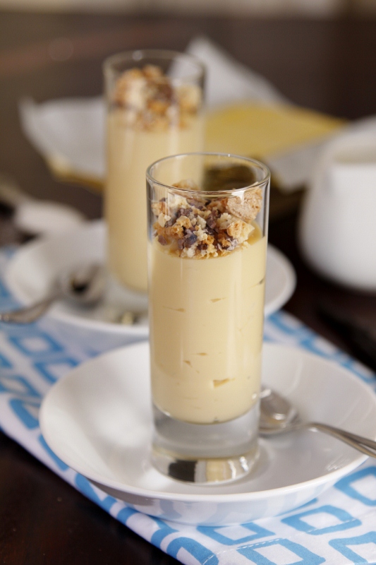 A glass of creamy dessert, inspired by Italian dessert recipes, topped with crumbled nuts or cookies, placed on a white plate with a spoon.