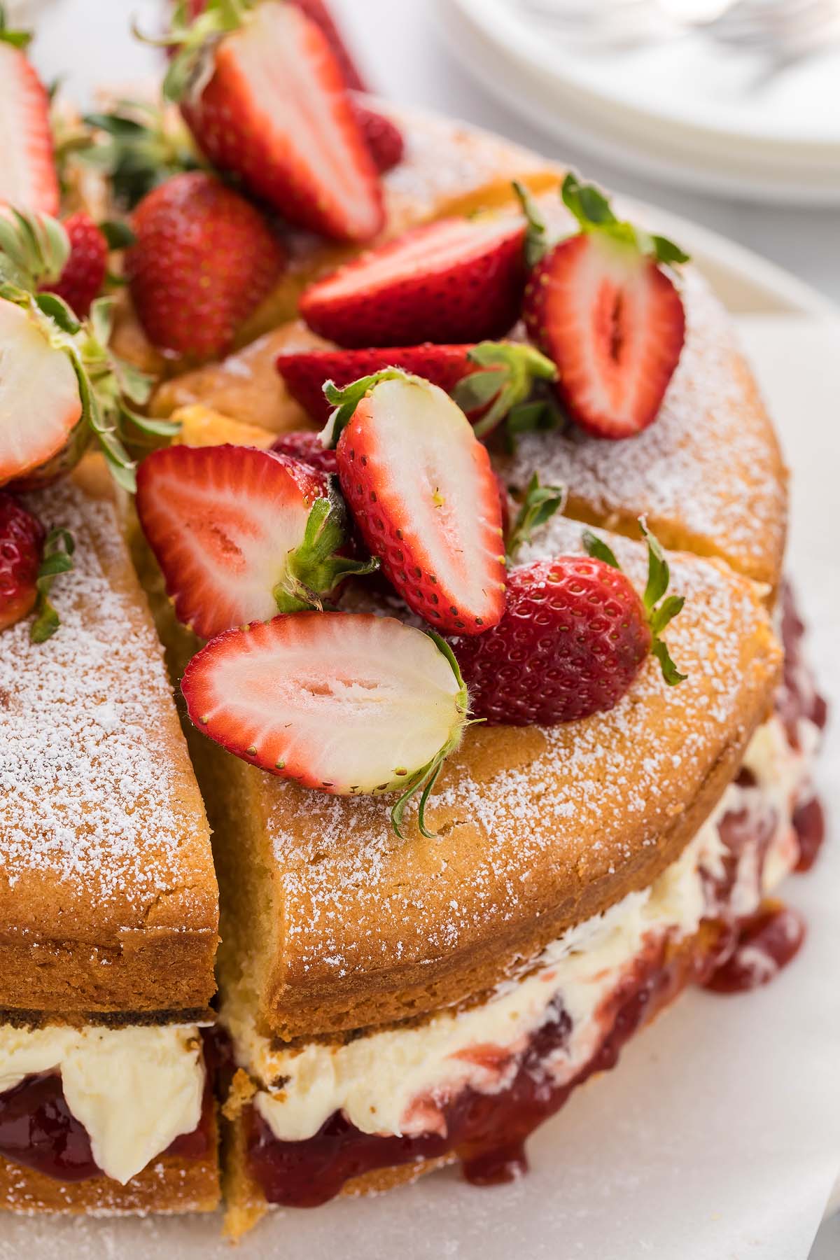 A classic Victoria Sandwich Cake featuring a sponge cake with cream and jam filling, topped with halved fresh strawberries and dusted with powdered sugar.