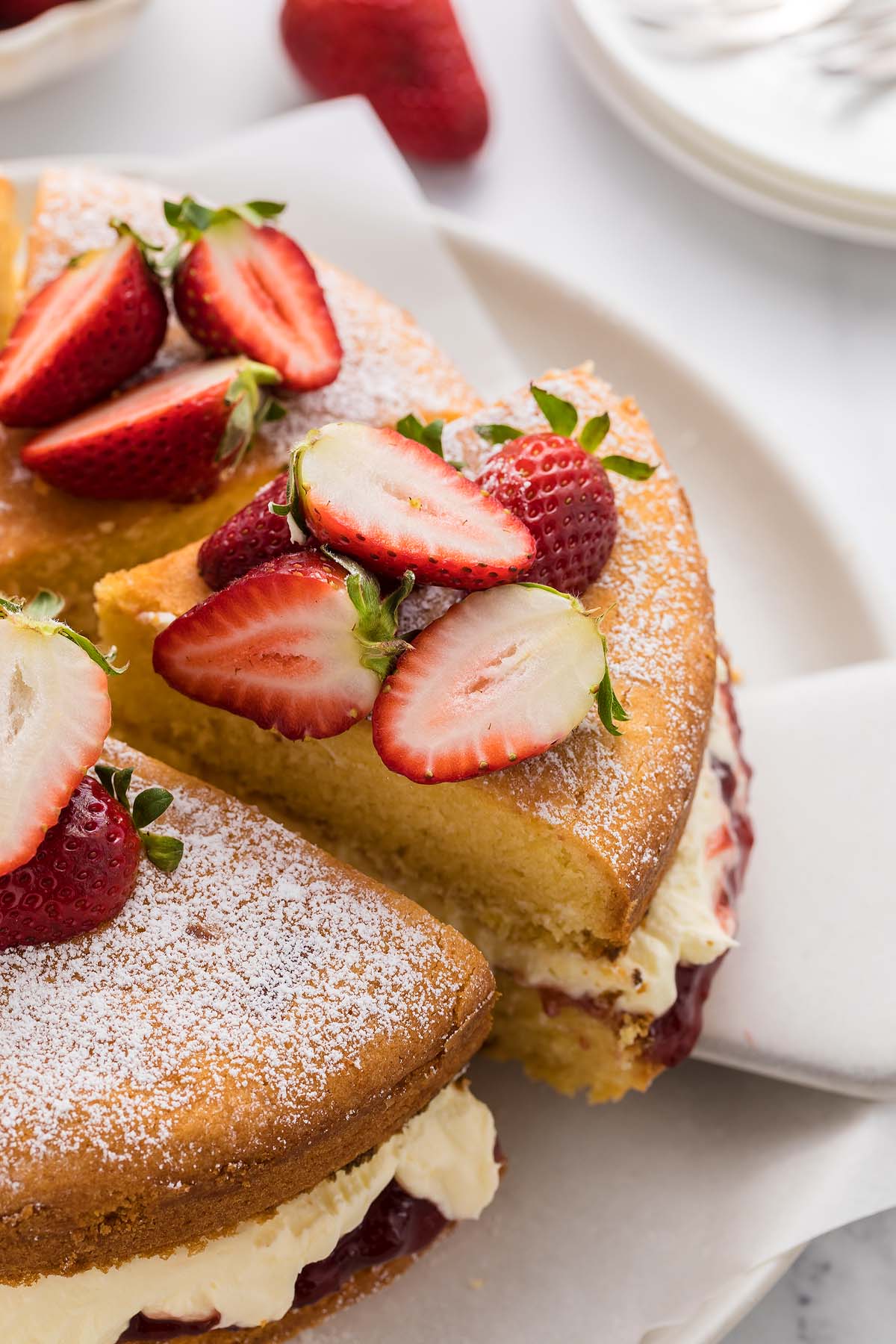 A classic Victoria Sandwich Cake: a slice of sponge cake with cream, jam, and fresh strawberries on top, dusted with powdered sugar.