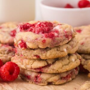 A stack of White Chocolate Raspberry Cookies sits on parchment paper, with fresh raspberries and a bowl in the background.