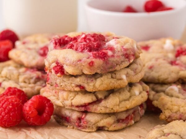 A stack of White Chocolate Raspberry Cookies sits on parchment paper, with fresh raspberries and a bowl in the background.