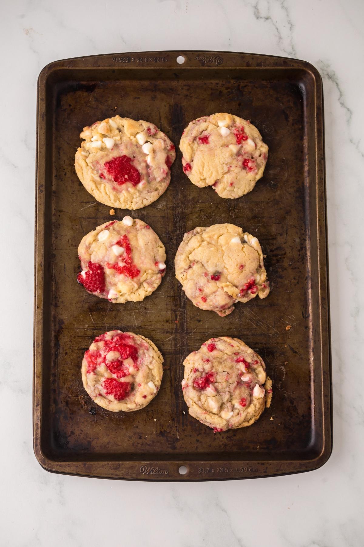 Six cookies with white chocolate chips and raspberries on a worn baking sheet, seen from above.