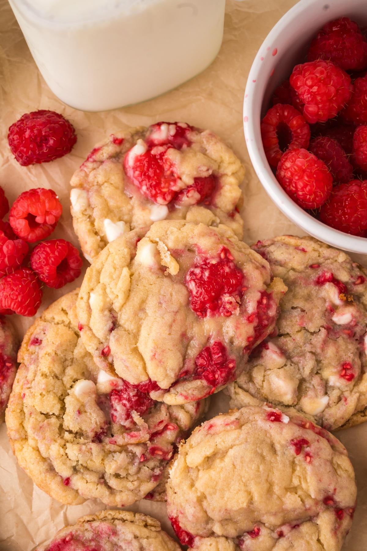 A close-up of raspberry cookies with white chocolate chips, fresh raspberries, and a glass of milk on parchment paper.