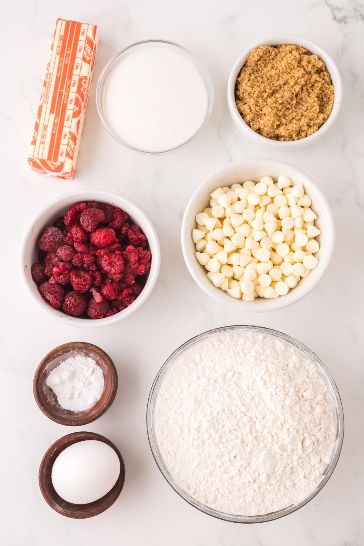 Ingredients on a white surface: butter, sugars, raspberries, white chocolate chips, flour, baking soda, and an egg.
