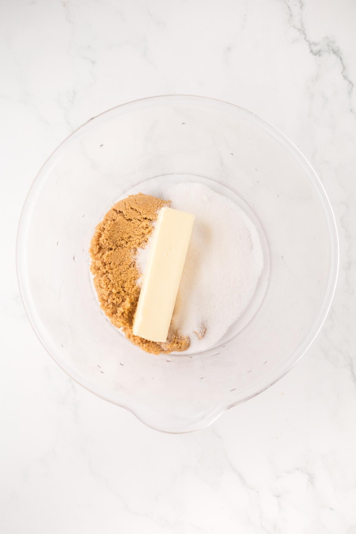 A glass bowl with a stick of butter, brown sugar, and white sugar on a white marble surface.