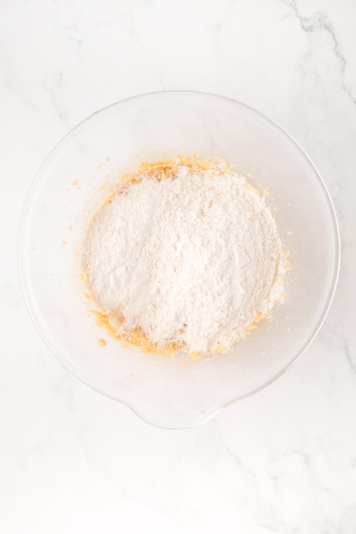 A glass bowl with flour and other baking ingredients on a white marble surface.