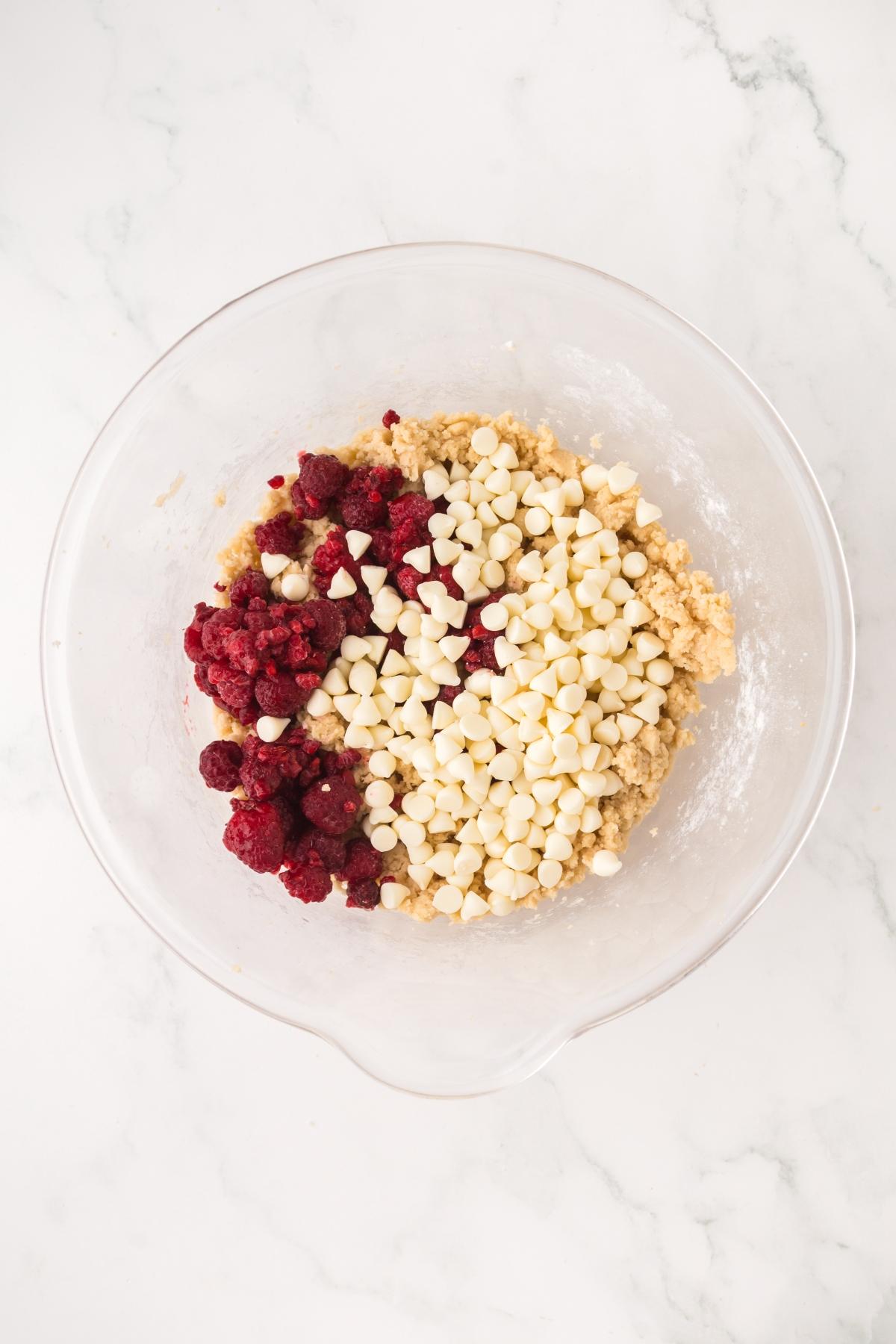 A glass bowl with cookie dough, white chocolate chips, and raspberries on a white marble surface.
