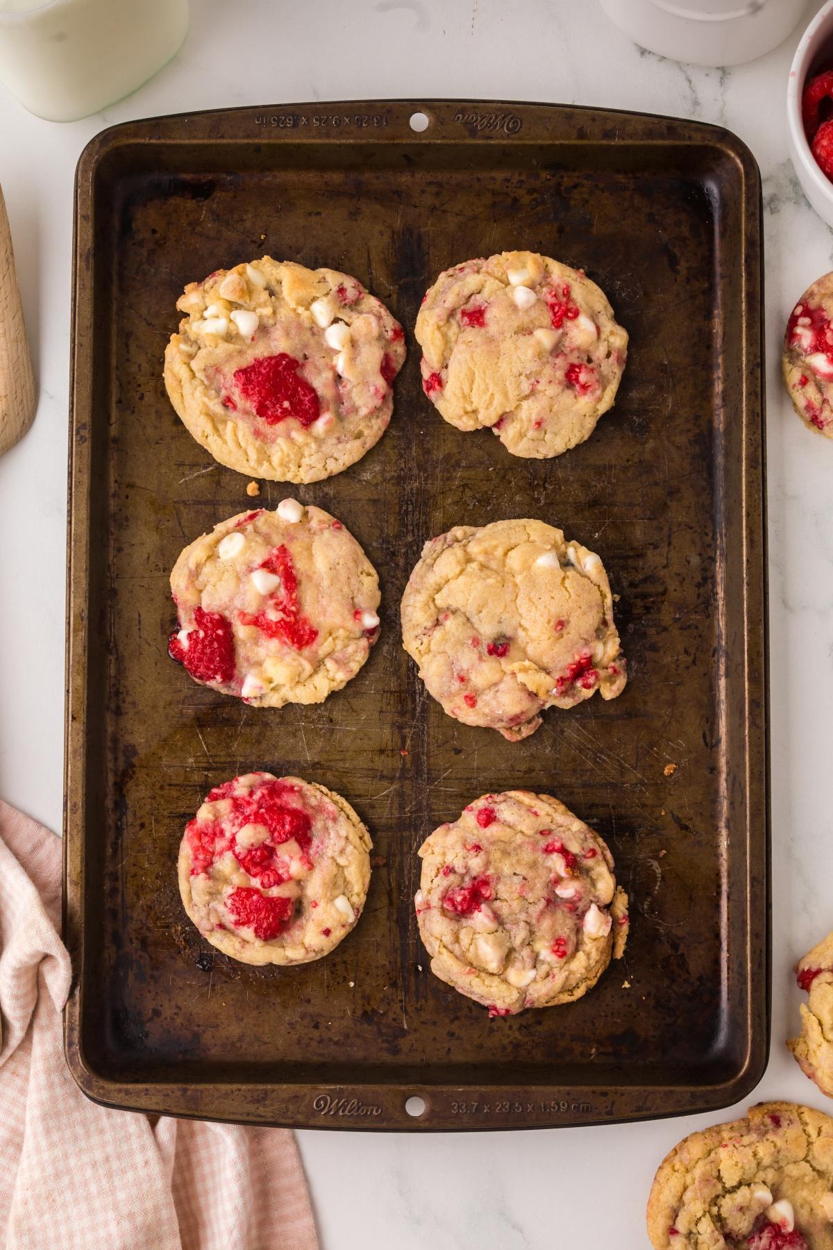 Six baked cookies with white chocolate chips and raspberries on a dark metal baking sheet.