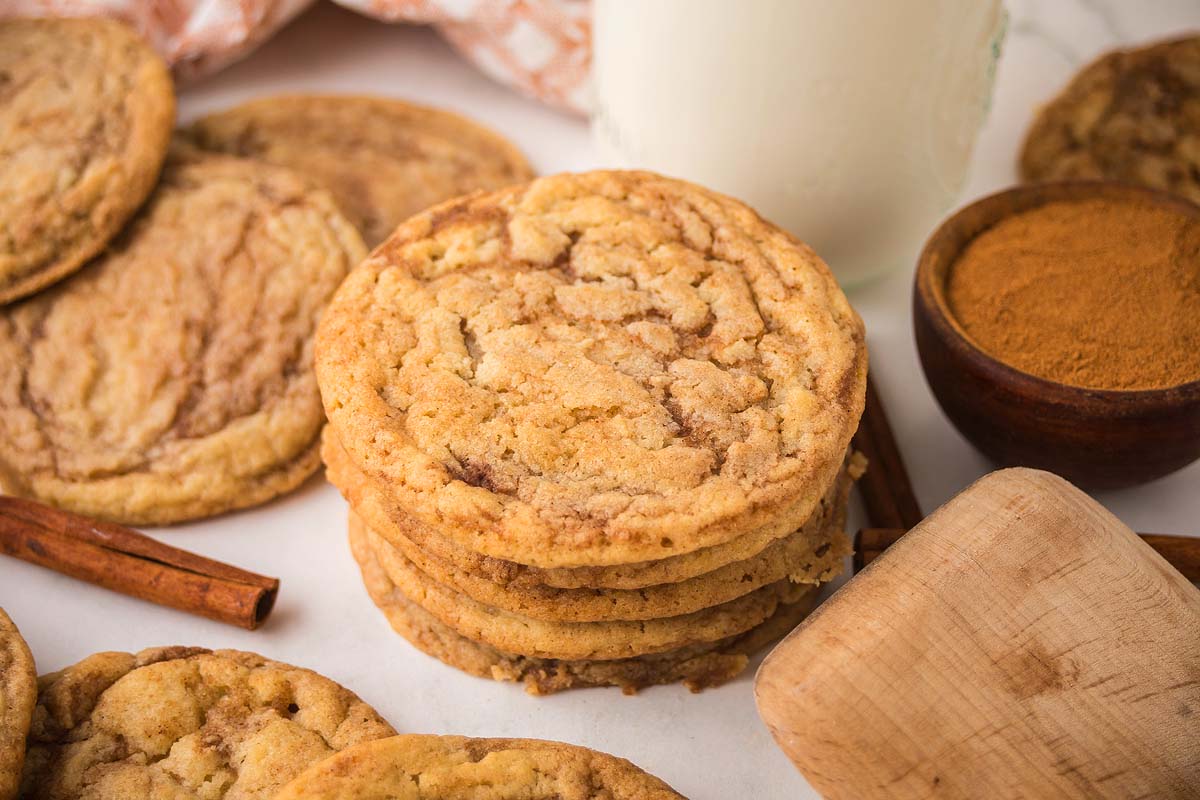A stack of cinnamon roll sugar cookies on a table with cinnamon sticks, a bowl of cinnamon, and a glass of milk.