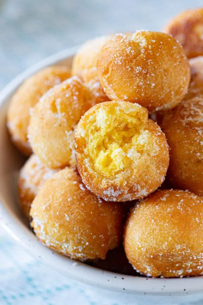 A bowl of sugared, golden donut holes inspired by Italian dessert recipes, with one showing a soft, fluffy inside.