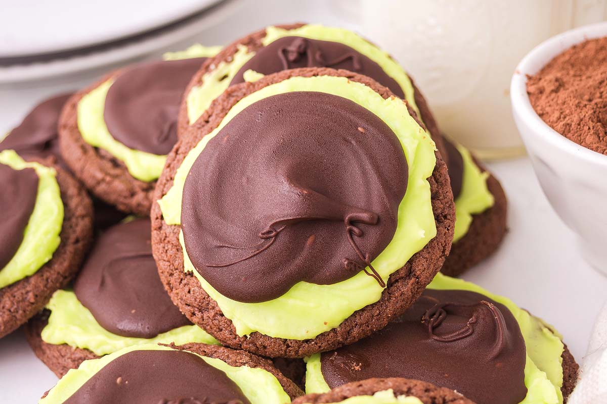 Chocolate mint cookies with green frosting and a layer of rich chocolate icing are stacked on a white surface.
