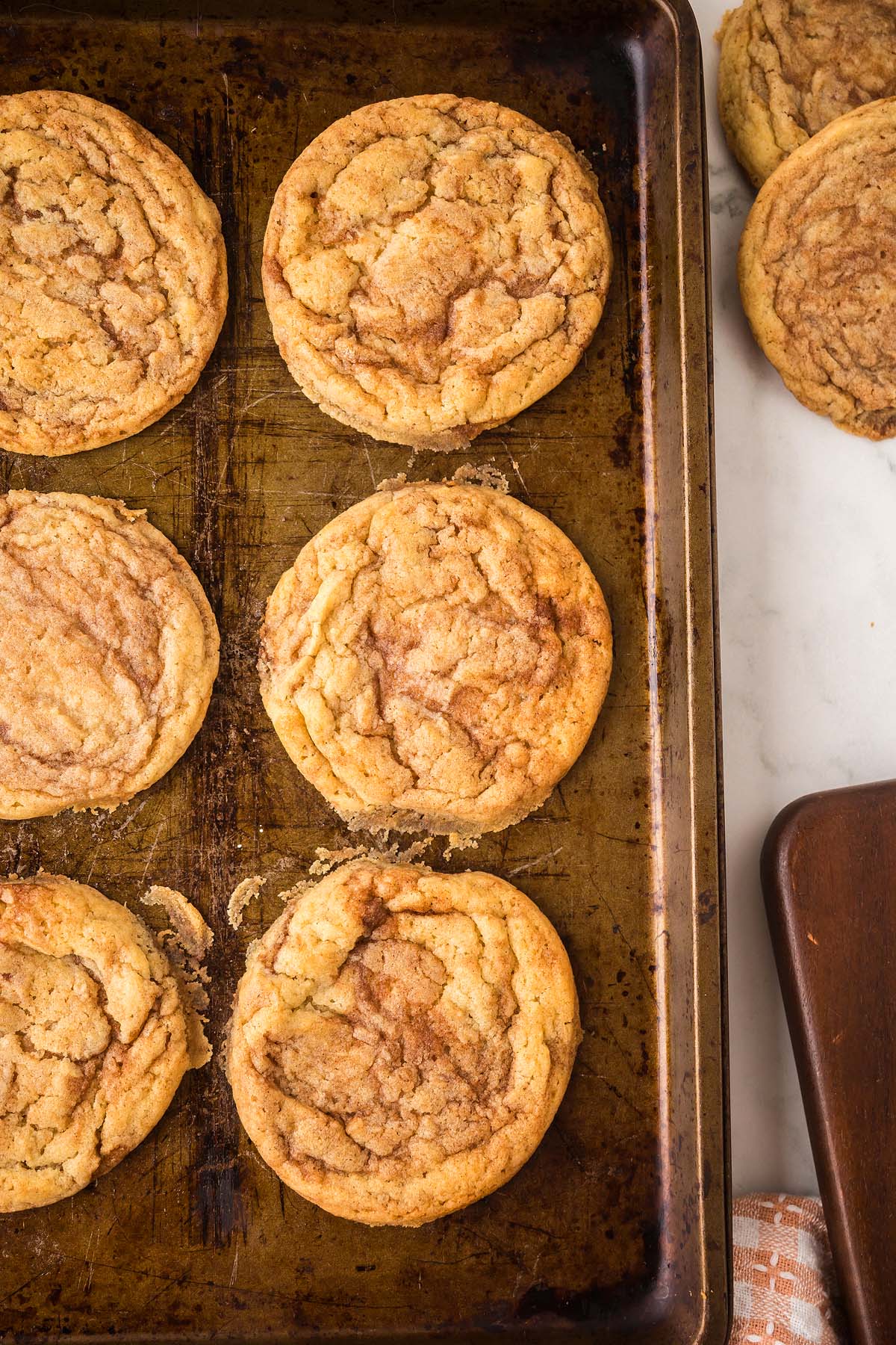Six cinnamon roll sugar cookies line a baking sheet, with two more cookies resting on a white surface nearby.