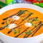 Bowl of tomato Caprese soup with cream, basil, and balsamic drizzle, surrounded by fresh vegetables and herbs.