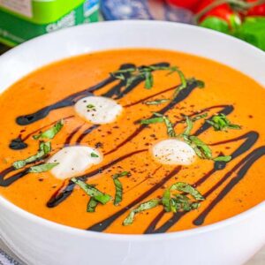 Bowl of tomato Caprese soup with cream, basil, and balsamic drizzle, surrounded by fresh vegetables and herbs.