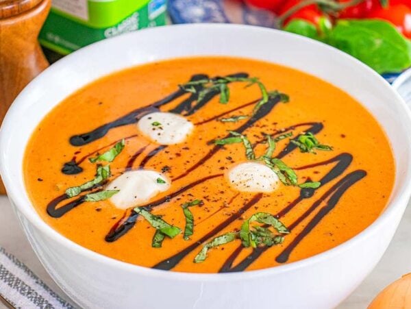 Bowl of tomato Caprese soup with cream, basil, and balsamic drizzle, surrounded by fresh vegetables and herbs.
