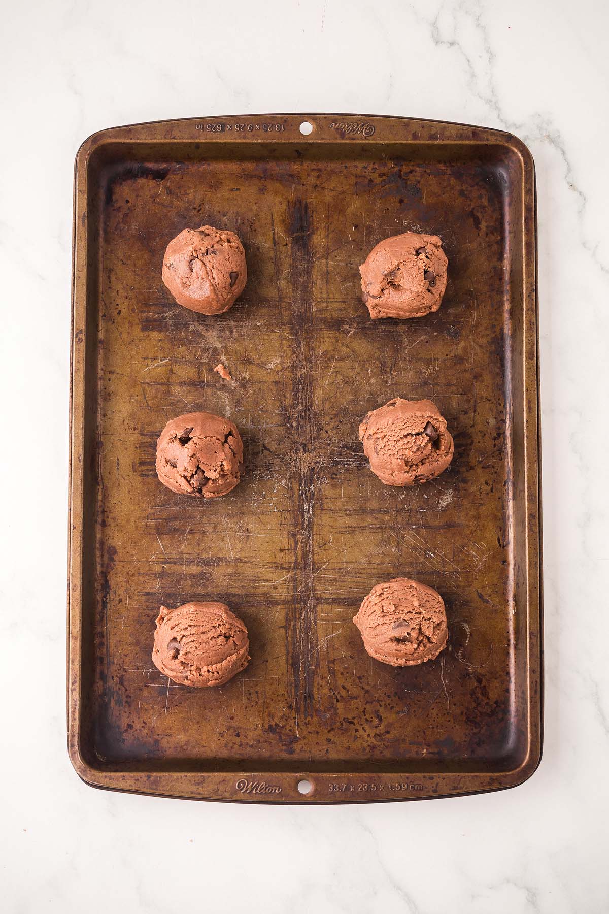 Six scoops of Chocolate Pudding Cookies dough are spaced evenly on a worn baking sheet, ready to be baked.